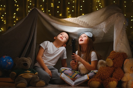 Dos niños pequeños juegan en casa por la noche para construir una tienda de campaña para leer libros con una linterna y dormir dentro. Concepto de: juego, magia, creatividad, sistemas de alarma. photo