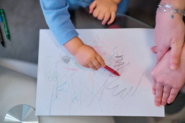 istock Toddler drawing on white paper with crayons while his mother helps him. 1452533295