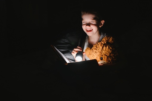 A boy of 5-6 years old is reading a book in the evening in the dark under a blanket with a toy bear