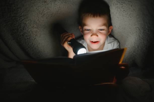 istock Boy with flashlight reading book under blanket at home. 1452536547