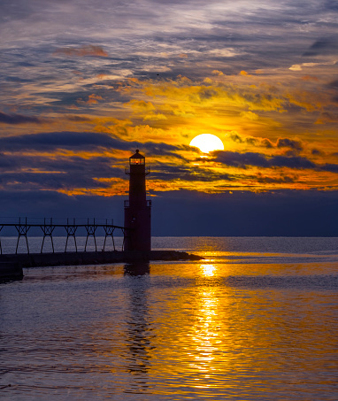 Vibrant textured sky and red lighthouse reflected in tranquil waters in harbor at sunrise.