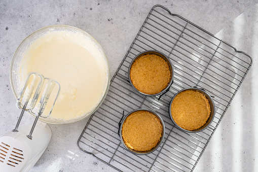 Cheesecake recipe. Three mini round cake pans (tin, mould), with crushed crackers, and cheesecake batter in a bowl, flat lay