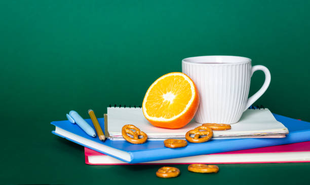 Notebooks, a cup of tea and an orange on a green background, the concept of a school snack, copy space.
