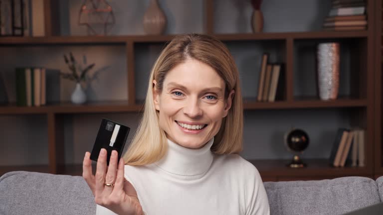 Blonde woman showing credit card. Smiling young customer holding bank card and laptop sitting on couch at home Happy female shopper using instant easy mobile payments making purchase in online store.