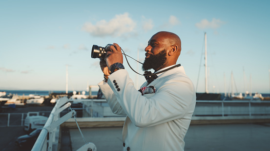 A profile view of a sophisticated bald African man with a well-groomed black beard, in a fashionable white suit, holding a camera with both hands while standing on a terrace at the marina