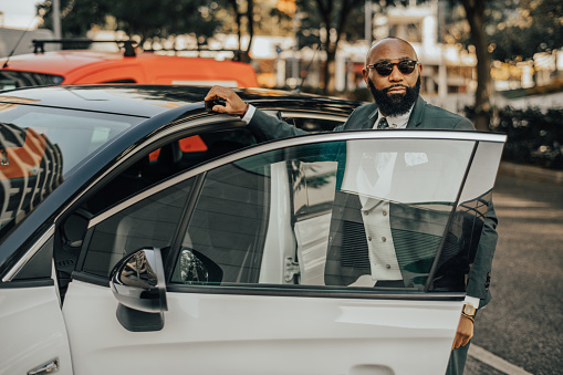 View of a sophisticated bald black businessman with a fine black beard, in dark sunglasses and a tailored fashionable suit, looking aside about to enter his white elegant car parked outdoors