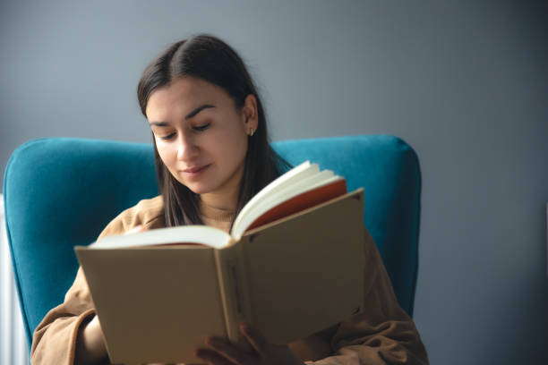 istock A young woman is reading a book while sitting in a chair. 1452563442
