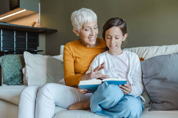 Grandmother reading a book to her granddaughter Grandmother and granddaughter are in the living room, grandmother is holding a book and reading short story stock pictures, royalty-free photos & images