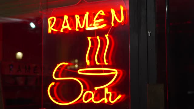 Video of a red neon lights that have the word ramen written next to a door of a city establishment. Letters on a sign indicating that ramen soup is for sale.