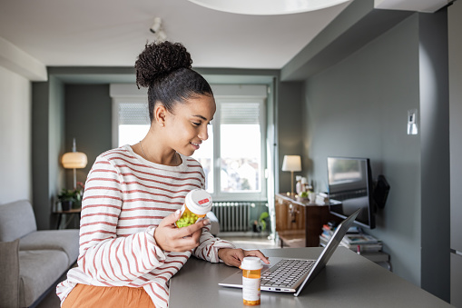 African-American woman holding a pill bottle while searching for the informations online. Technology makes life easier.