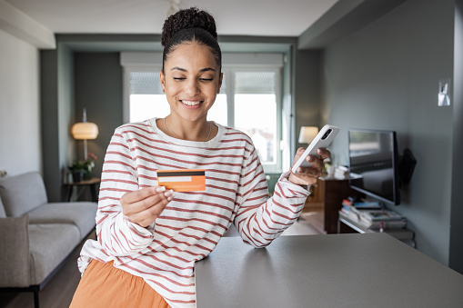 Young African-American woman at home shopping online. She is holding mobile phone and credit card. E-banking via mobile app.