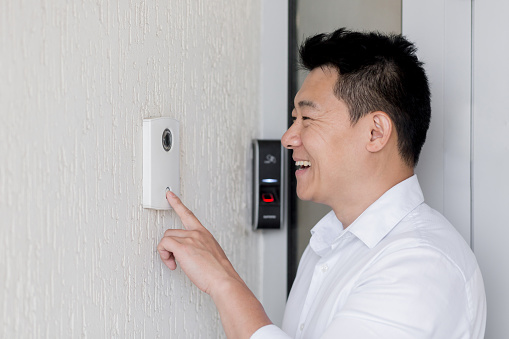 Close-up photo. A young Asian man rings an electronic doorbell on the wall of a house with his finger. He opens the door to his home.