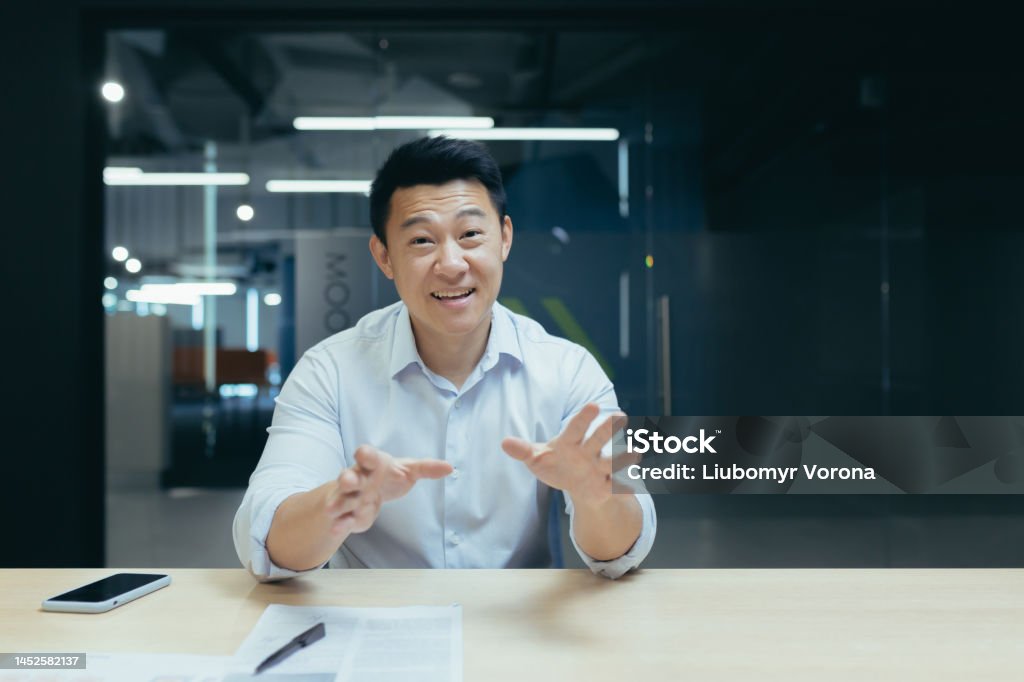 An Asian man sitting in the office and talking to the camera. Online meetings, business training Smiling young Asian man sitting in the office at the desk and talking to the camera. Conducts online meetings, consults, business training. Discussion Stock Photo