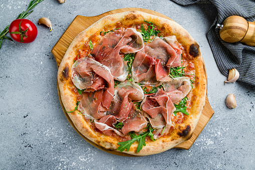 Pizza with Parma ham and arugula on wooden board on grey table top view