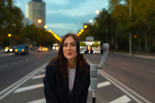 Smiling young woman with long hair in warm clothes standing on traffic island and taking self portrait via cellphone placed on tripod