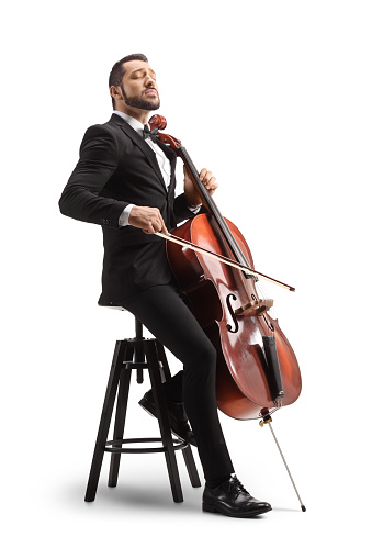 Male musician in a black suit and bow-tie sitting on a chair and playing a cello isolated on white background