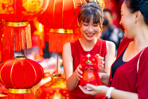 Happy Asian family mother and daughter holding red shopping bag during choosing and buying home decorative ornaments and joss paper for celebrating Chinese Lunar New Year festival at Chinatown street market. Chinese culture concept.