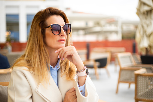 Beautiful blond woman sitting and relaxing outdoors. Woman is wearing white coat, blue shirt and sunglasses and a smart watch, looking into the distance and relaxing. Background is out of focus, a modern hotel terrace.