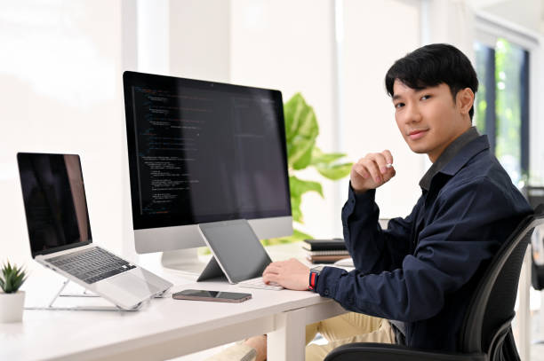 istock Smart Asian male web developer sits at his modern office desk, smiling and looking at the camera. 1452609778