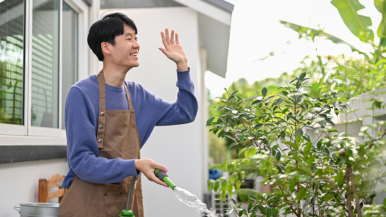 Happy and cheerful young Asian man waving hand, greeting his neighbor while watering his plants at the backyard.