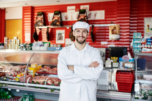 ein metzgerverkäufer steht mit verschränkten armen in der metzgerei und lächelt in die kamera. - supermarket meat store manager stock-fotos und bilder