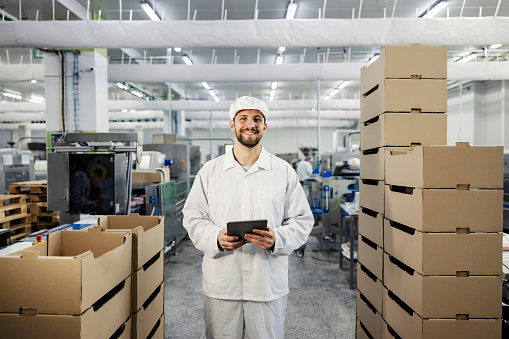 A happy meat industry supervisor stands between the boxes in a factory with a tablet in his hands and smiles at the camera.