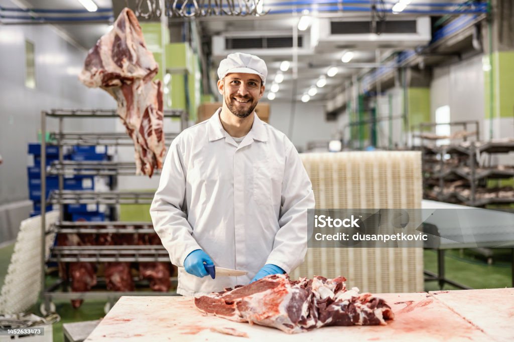 A butcher is working with raw meat in meat factory. A butcher is cutting raw meat into pieces while smiling at the camera. Butcher Stock Photo