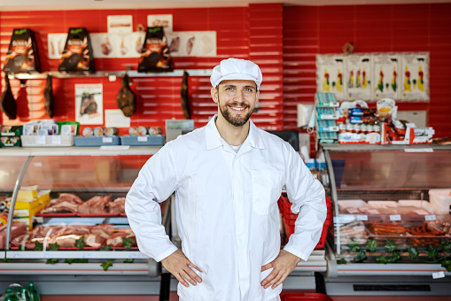 A successful meat shop worker is standing in a shop and smiling at the camera.