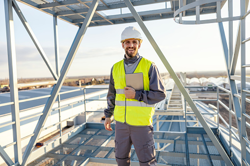 An industry worker is standing on silo with tablet in hands and smiling at the camera.