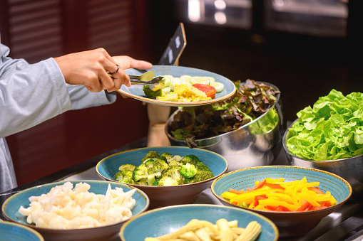 A guest in a restaurant pours fresh vegetables into his plate, salad bowls are full on the buffet counter