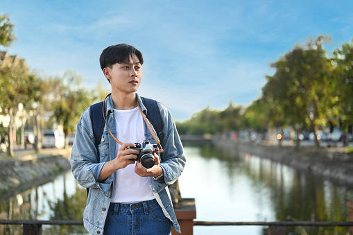 Young asian male blogger with digital camera standing on bridge with scenery view of river and beautiful blue sky.