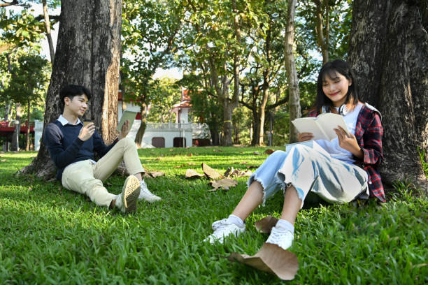 Two smiling students reading books, preparing for exam while under tree at sunny beautiful day. Education and lifestyle concept.