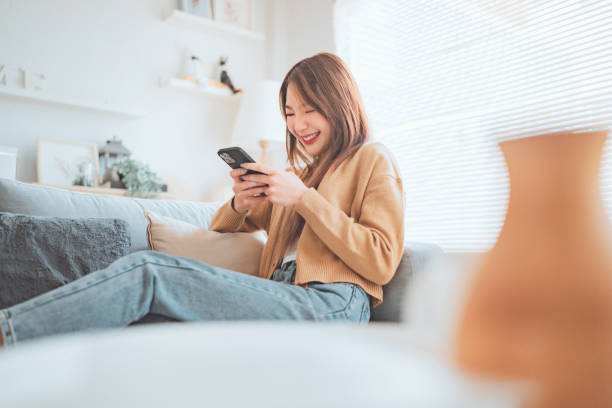 istock Happy young asian woman browsing surfing wireless internet on mobile phone while sitting a couch in living room at home. 1452648195