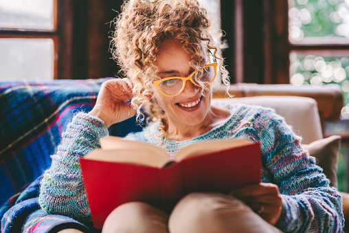 Serene people enjoying relax and indoor leisure activity at home reading a book with red cover. Education concept lifestyle. One happy female people read novel sitting at home. Smiling lady enjoying