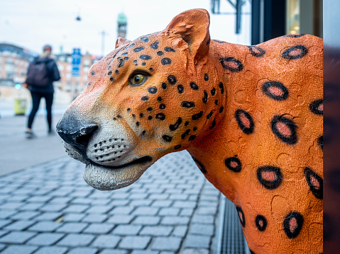 Orange cheetah figurine head sticking out toward the pavement with a curious look. Pavement and pedestrian behind (selective focus).