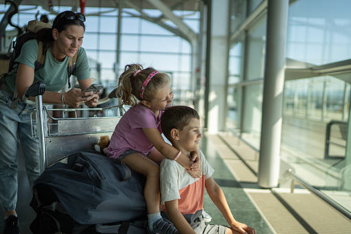 Little brother and sister sitting and playing on luggage cart while mother texting on mobile phone on the airport