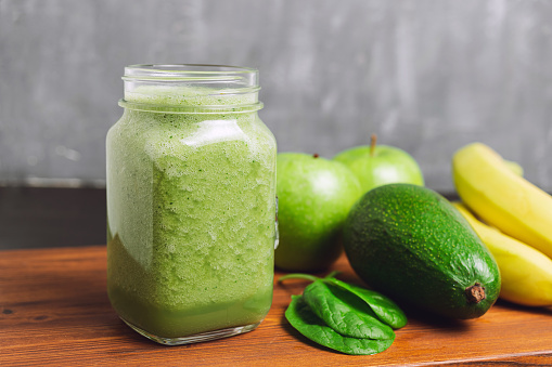 Green smoothie drink blended in a glass jar, avocado, spinach leaves, banana and apples at wooden board.
