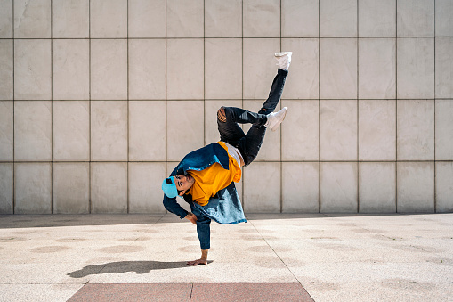 Talented young man doing break dance dances against white wall in the street and looking at camera.