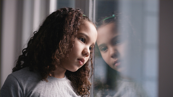 Window, depression and children with a bored girl by a transparent glass door in her home during winter. Thinking, kids and alone with a sad female child looking out from inside of a house