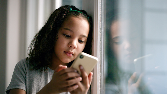istock Window, smartphone and girl on social media, connection and search internet in house. Young female, kid and child with phone, online reading or communication being calm, playing games, device or chat 1452661352