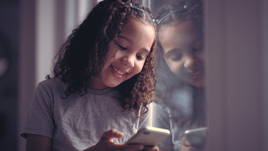 Social media, chat and child with phone for communication, web games and cartoon with a window reflection in a house. Education, learning and girl with a mobile for app, relax and online conversation