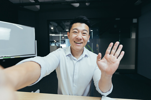 Portrait of a young handsome Asian man taking a selfie, recording a video, talking on a video call, holding a phone, waving and greeting the camera, smiling.