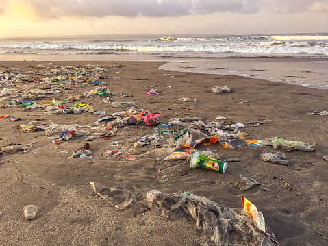 Full frame shot of heap plastic waste lying on the ground covering the sand of a beautiful beach in Kuta, Bali, during sunrise. In the background some unrecognizable surfer walking on the dirty beach