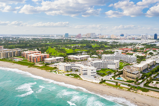 Aerial drone view of palm beach area, with hotels and buildings, beach, green areas, tropical vegetation, turquoise sea, waves, sand, blue sky