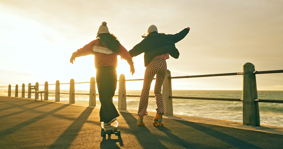 Skate, sunset and back of friends by the beach on a board and skates for happiness, fun and sport. Skateboard, rollerskate and cool women with fashion, urban and young style while skating by the sea