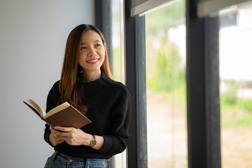 Asian woman standing near window and reading book at home.