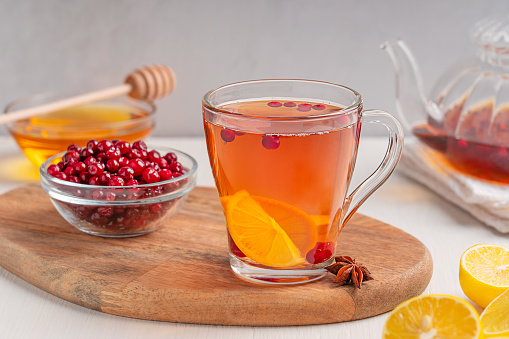 Homemade healthy organic antioxidant hot tea drink served in transparent glass cup with red cranberries, lemon citrus slices and honey on white wooden table with teapot and ingredients at kitchen
