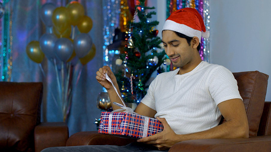 Attractive Indian guy happily opens a gift box while sitting near a decorated Christmas tree during Christmas season