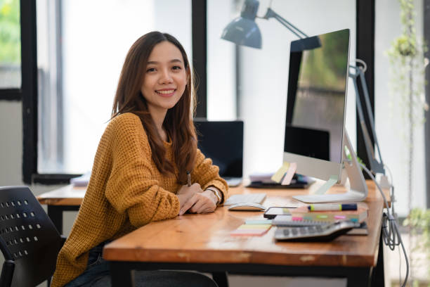istock Portrait of a Beautiful Asian freelancer Using Desktop Computer at the home office. Smiling and looking at the camera. 1452684396