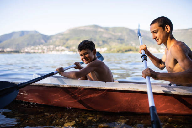 A couple paddles a red canoe on a serene lake, surrounded by lush, forested hills. The calm water reflects the clear blue sky, capturing the tranquility of nature and a sense of peaceful solitude.